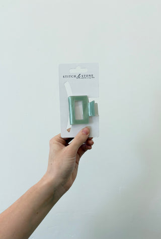 A hand holding a square-shaped green and beige hair clip, packaged in a white plastic container with the brand 'Stitch & Stone' visible on it.