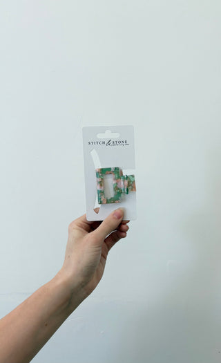 A hand holding a square-shaped green and beige hair clip, packaged in a white plastic container with the brand 'Stitch & Stone' visible on it.