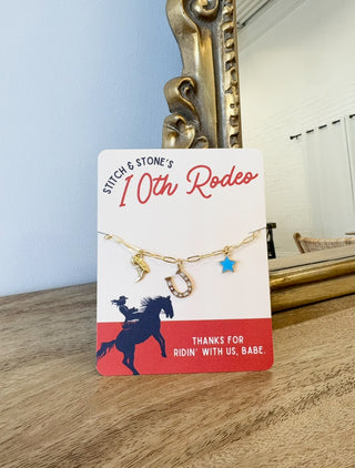 Gold necklace with charms on a '10th Rodeo' card in front of a decorative mirror.