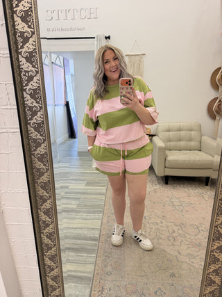 Person taking a mirror selfie wearing a pink and green outfit in a room with a large mirror.