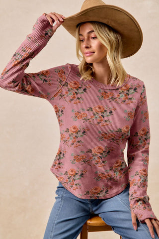 Woman wearing a pink floral long-sleeve top and blue jeans, sitting on a beige background.