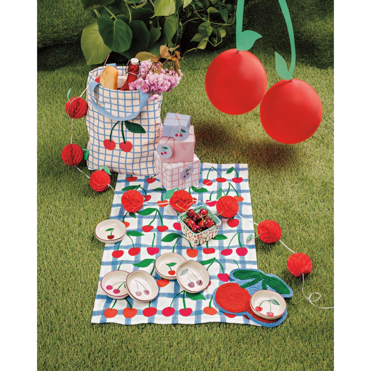 Picnic setup with a checkered bag, plates, and cherry-themed decorations on grass.