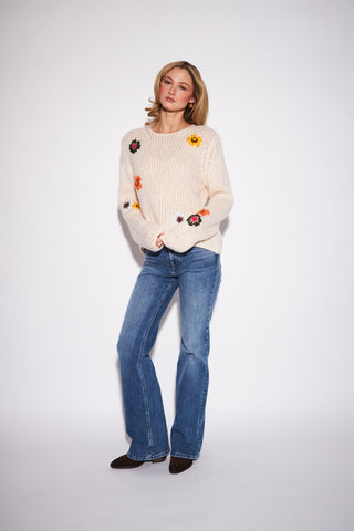 A woman modeling a cream-colored chunky knit sweater with colorful embroidered flowers, paired with blue jeans and dark brown shoes, against a plain white studio background.