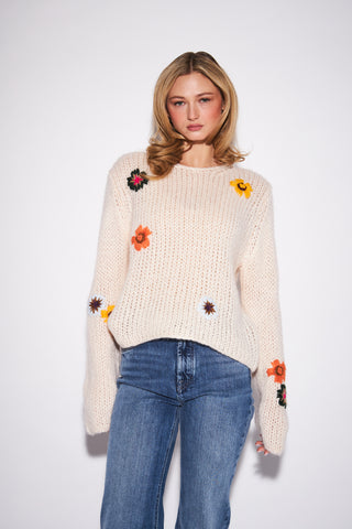 A woman modeling a cream-colored chunky knit sweater with colorful embroidered flowers, paired with blue jeans, against a plain white studio background.