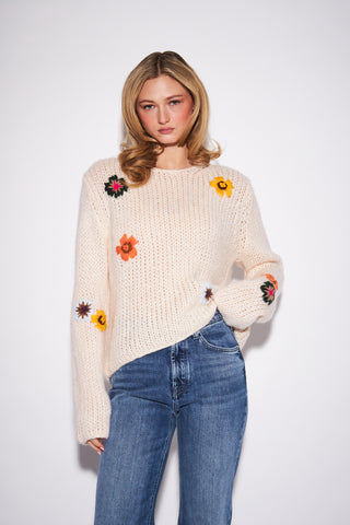 A woman modeling a cream-colored chunky knit sweater with colorful embroidered flowers, paired with blue jeans, against a plain light gray background.
