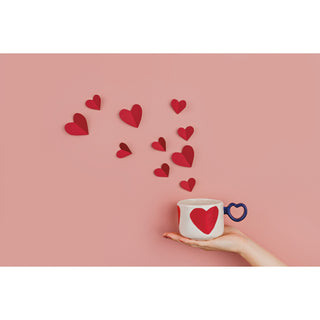 Hand holding a mug with a heart design, surrounded by scattered red hearts on a pink background