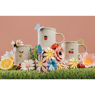 Three ceramic mugs with floral and fruit designs on a grassy surface with flowers and a lemon.