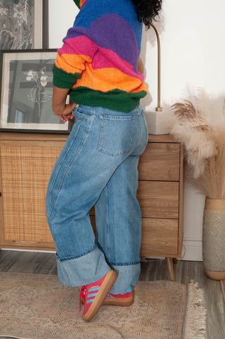 Person wearing a colorful sweater and blue jeans in a room with wooden furniture and decorative items.