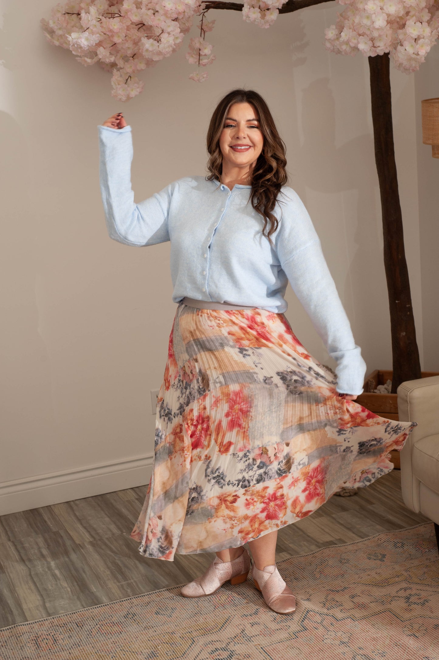 Woman in light blue shirt and floral skirt standing indoors with cherry blossom branches in the background.