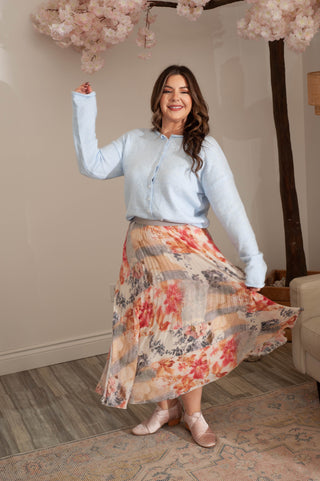 Woman in light blue shirt and floral skirt standing indoors with cherry blossom branches in the background.