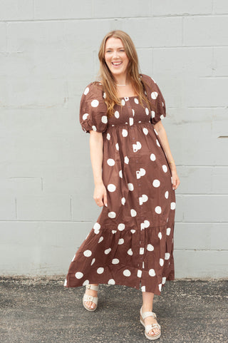 A woman modeling a brown midi dress with white polka dots, standing against a light gray painted brick wall.