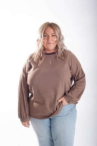 A woman modeling a brown long-sleeve crewneck top with fine horizontal stripes, paired with light blue jeans, against a plain white studio background.