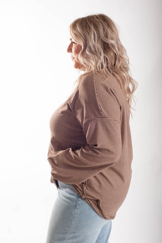 A woman in profile wearing a brown and beige vertically striped long-sleeve top and light blue jeans, photographed against a plain white studio background.