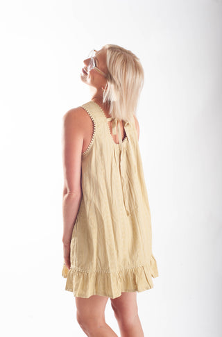 A woman wearing a sleeveless light beige linen dress with a ruffled hem and tie-back detail, photographed from the back against a plain white studio background.