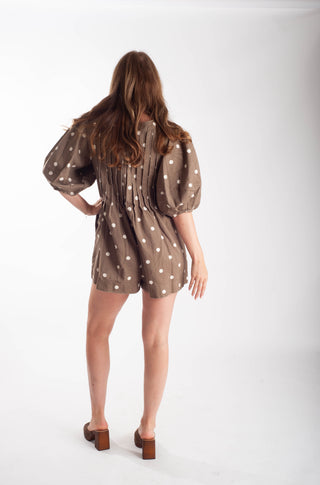 A woman viewed from the back wearing a brown short romper with white polka dots and puffed sleeves, paired with brown block-heel sandals, against a plain white studio background.