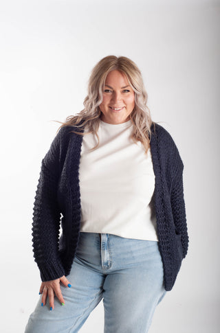 A woman modeling a dark navy blue chunky knit cardigan over a white top and light blue jeans, photographed against a plain light gray studio background.