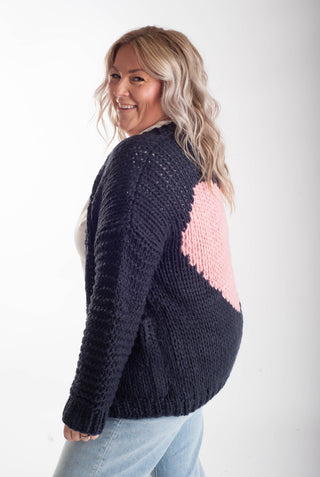 A woman wearing a chunky knit cardigan with a dark navy blue body and a large light pink heart on the back, paired with light blue jeans against a plain white studio background.