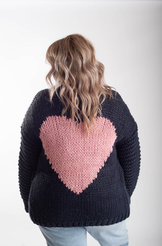 A woman viewed from behind wearing a chunky black knit sweater with a large pink heart on the back, paired with light blue jeans, against a plain white studio background.