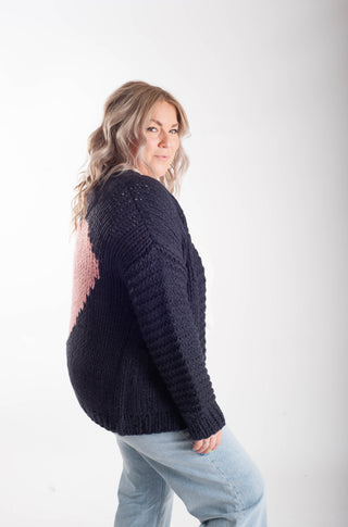 A woman wearing a chunky knit navy sweater with a pink textured heart design on the back, paired with light blue jeans, photographed against a plain white studio background.