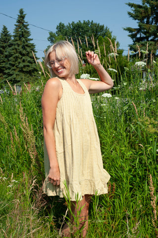 A woman wearing a sleeveless, light beige pleated dress stands in a grassy field outdoors under a blue sky.