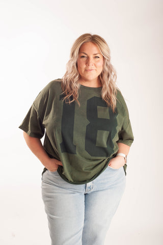 A woman modeling an oversized olive green t-shirt with a large dark green number 18 graphic on the front, paired with light-wash blue jeans, against a plain white studio background.