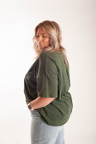 A woman with blonde hair is shown from a side-back angle wearing an oversized olive green short-sleeve t-shirt and light blue jeans against a plain white studio background.