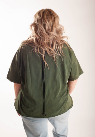 A woman modeling an oversized olive green t-shirt with a large dark green number 18 graphic on the front, paired with light-wash blue jeans, against a plain white studio background.