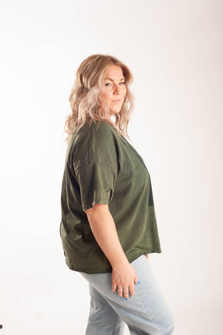 A woman wearing an oversized olive green short-sleeve top and light blue jeans, standing against a plain white studio background.