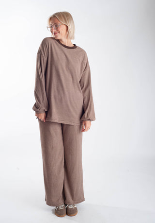 A woman modeling a matching two-piece lounge set consisting of a long-sleeve cropped top and wide-leg pants in a textured taupe-brown fabric, paired with brown shoes, against a plain white studio background.