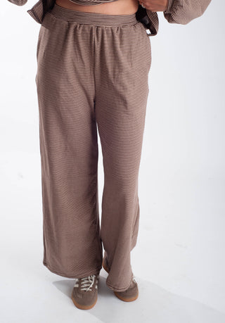 A person wearing loose, wide-leg pants in a brown and beige striped fabric, paired with brown shoes, shown from the waist down against a plain white studio background.