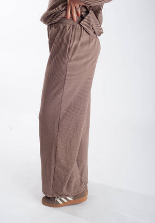 A person wearing loose, wide-leg brown pants with thin vertical stripes, paired with a matching top and brown slip-on shoes, shown from the waist down against a plain white studio background.