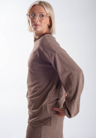 A woman with short blonde hair and glasses, wearing a brown and beige horizontally striped long-sleeve top, photographed from the side against a light gray studio background.