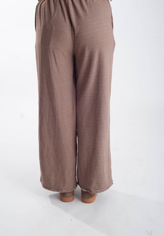 A person shown from the back wearing loose, wide-leg pants in a muted taupe-brown color with fine horizontal stripes, standing against a plain white studio background.