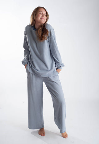 A woman modeling a matching two-piece lounge set consisting of a long-sleeve top and wide-leg pants in a dusty slate blue color, standing against a plain white studio background.