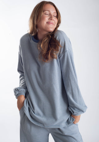 A woman wearing a matching long-sleeve striped lounge set in a dusty blue-gray and white color combination, standing against a plain light gray background.