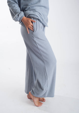 A person wearing a matching long-sleeve top and wide-leg pants set in a dusty blue-gray color with fine horizontal stripes, standing barefoot against a white studio background.