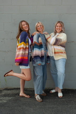 Three women standing together against a gray painted brick wall, each wearing a chunky knit striped cardigan in different colorways. The cardigans feature horizontal stripes in various combinations including purple, blue, pink, orange, yellow, and cream.