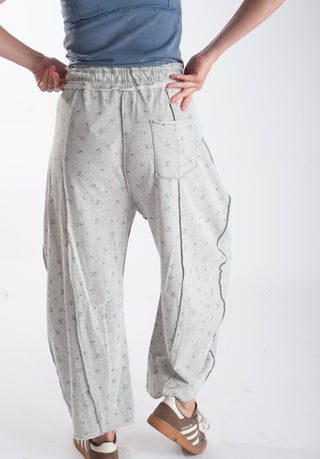 A person wearing light gray patterned lounge pants with an elastic waistband, side pockets, and a small floral print, paired with a blue top and brown sneakers, shown against a plain white studio background.