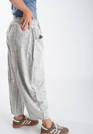 A person wearing light gray wide-leg pants with a small blue floral pattern, paired with a blue shirt and brown sneakers, shown from the waist down against a white studio background.