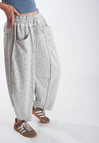 A person wearing loose, baggy light gray pants with a small allover floral print, side pockets, and an elastic drawstring waist, paired with brown sneakers against a plain white studio background.