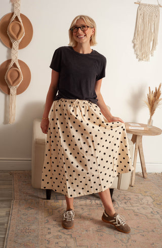 Woman wearing a black t-shirt and beige polka dot skirt in a room with decorative items.