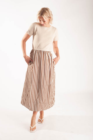 A woman modeling a beige short-sleeve top and a brown and white vertically striped midi skirt with side pockets, paired with white flat sandals, against a plain white studio background.