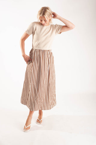 A woman modeling a beige short-sleeve top and a brown and beige vertically striped midi skirt, paired with white thong sandals, against a plain white studio background.