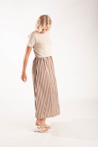 A woman wearing a cream short-sleeve top and a brown and white vertically striped midi skirt, standing against a plain white studio background.