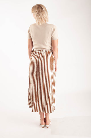 A woman viewed from behind wearing a beige short-sleeve top and a brown and beige vertically striped midi skirt, paired with white pointed-toe flats, against a plain white studio background.