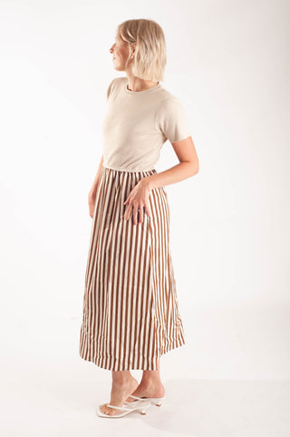 A woman modeling a beige short-sleeve crewneck t-shirt and a brown and beige vertically striped midi skirt, paired with white slide sandals, against a plain white studio background.