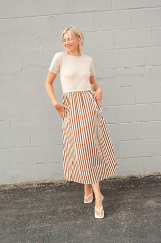 A woman stands against a light gray painted brick wall wearing a cream short-sleeve knit top and a knee-length skirt with vertical brown and cream stripes. She is wearing cream wedge sandals.