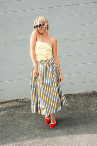 A woman wearing a pale yellow strapless top and a beige-and-white vertically striped midi skirt, paired with red pointed-toe heels and dark sunglasses, standing against a light gray brick wall.