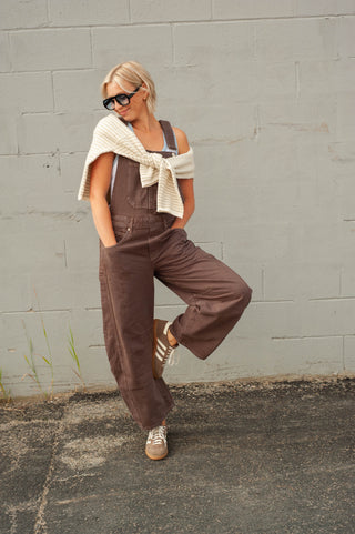 A woman posing in brown wide-leg denim overalls with a cream knit sweater draped off her shoulders, standing against a gray brick wall.