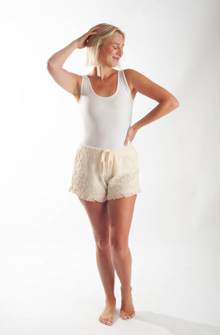 A woman modeling a white ribbed tank top and cream lace shorts against a plain white studio background.